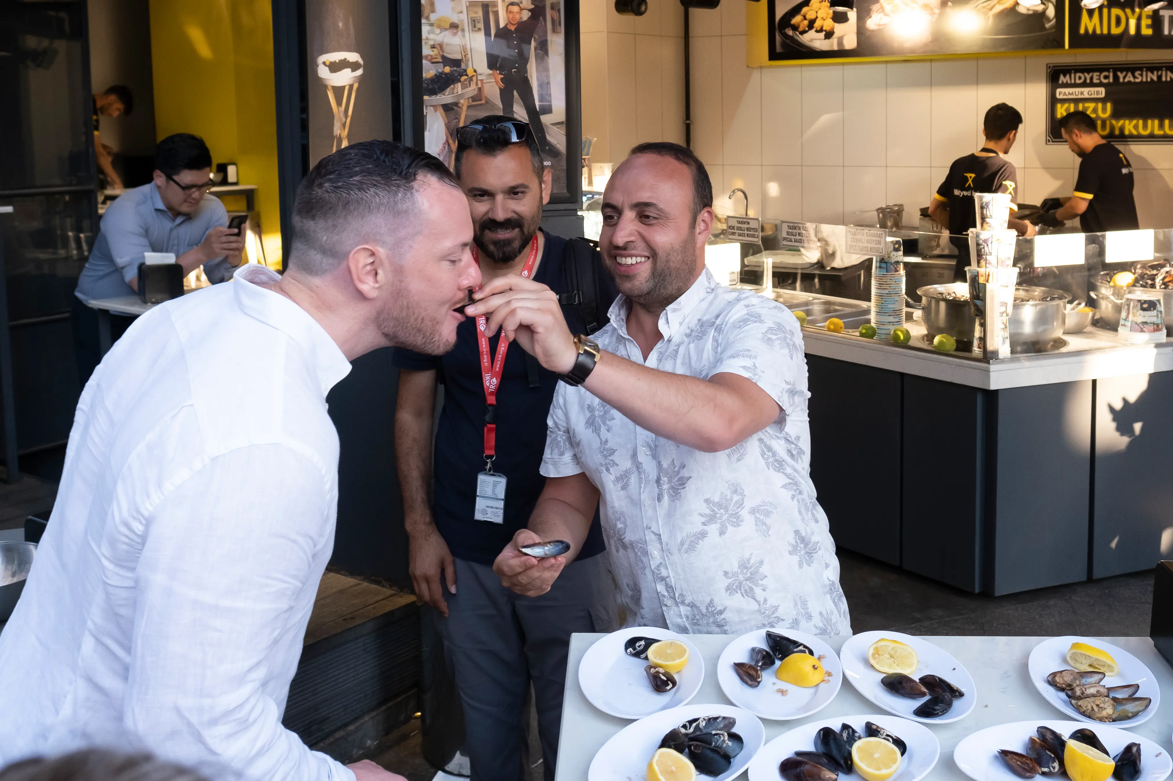 A guide feeds a guest a stuffed mussel at Midyeci in Istanbul while another guest watches and laughs