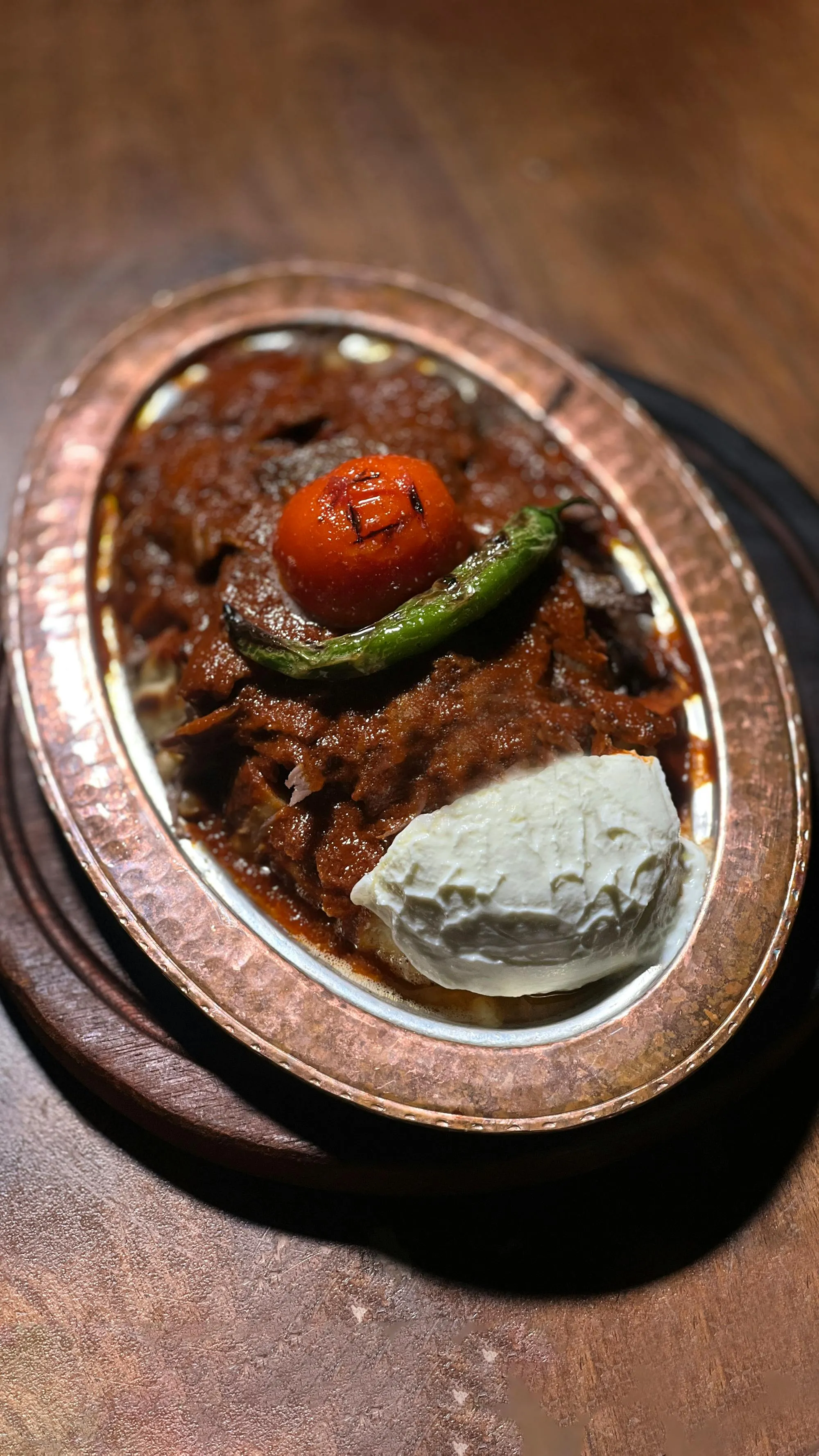 A traditional İskender kebab plate: sliced döner on cut-up flatbread under tomato sauce, topped with a roasted tomato and green pepper, yogurt on the side, in a hammered copper bowl
