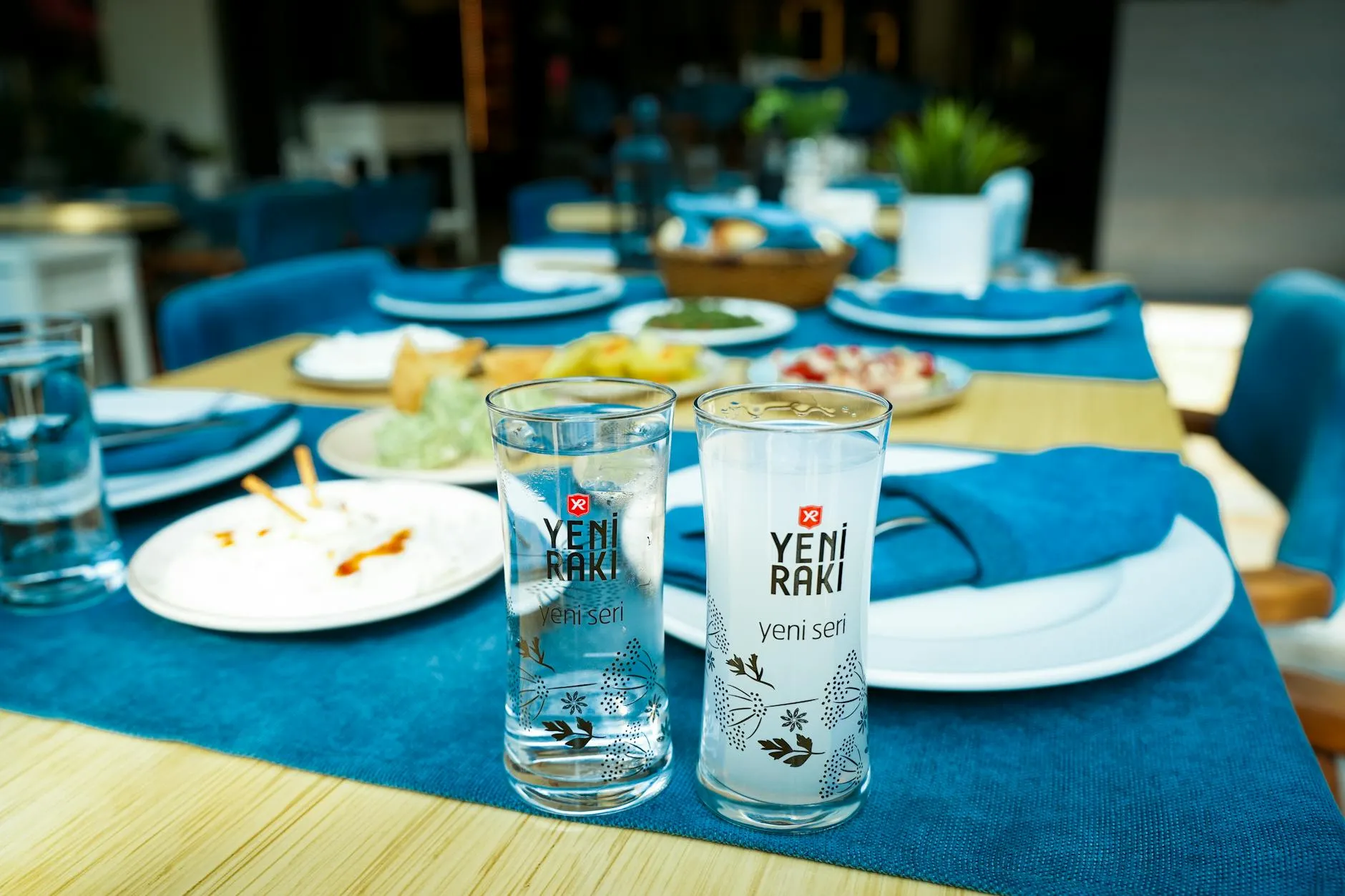 A classic meyhane table set with two tall Yeni Rakı glasses, several small meze plates including dips, and breadsticks on a blue tablecloth