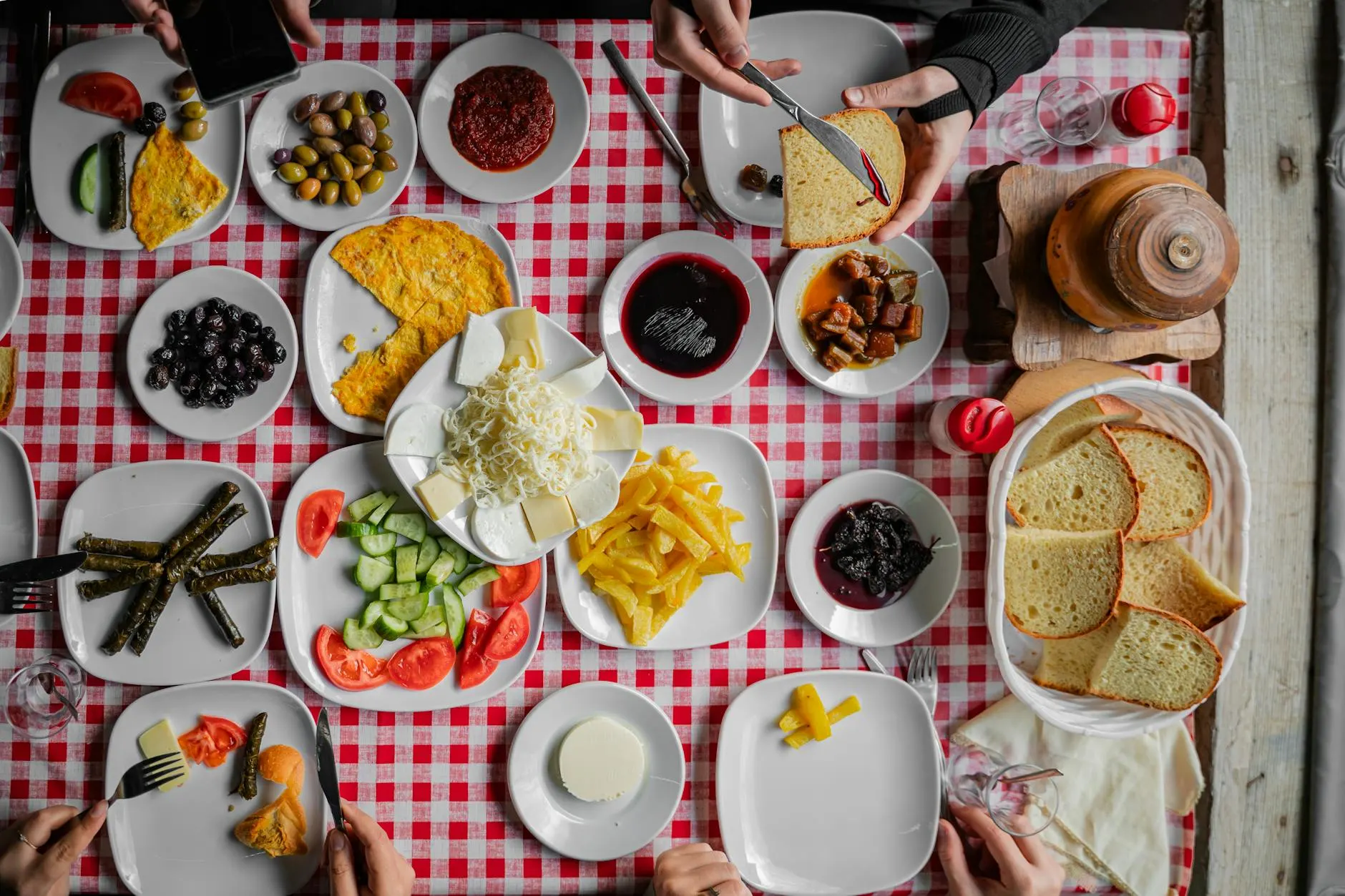 An overhead Turkish serpme kahvaltı: cheeses, olives, eggs, cucumber, tomato, bread, jams and tea laid out across a red-checkered table