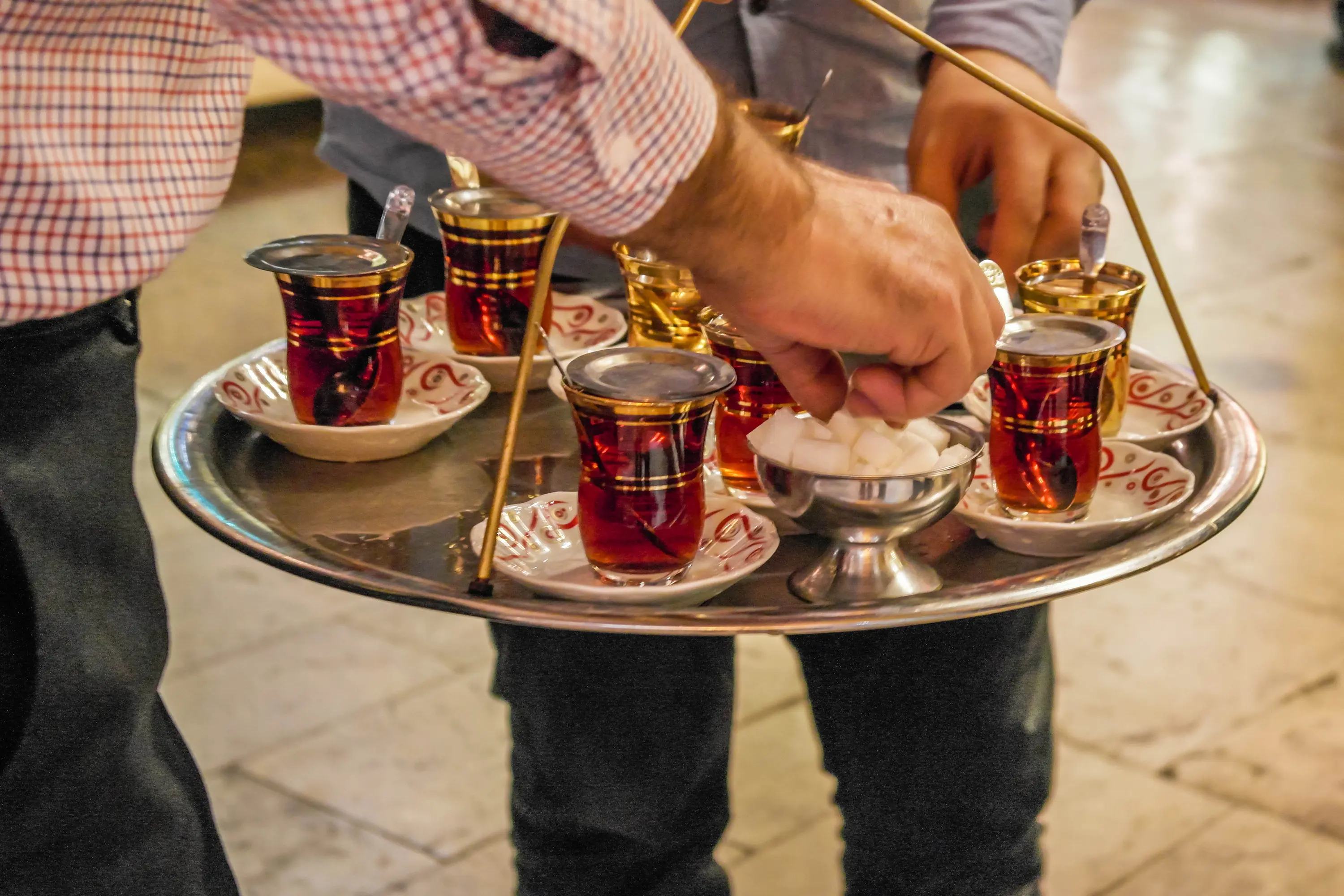 A waiter carrying a round metal tray of tulip-shaped Turkish tea glasses, reaching for the sugar bowl in the middle
