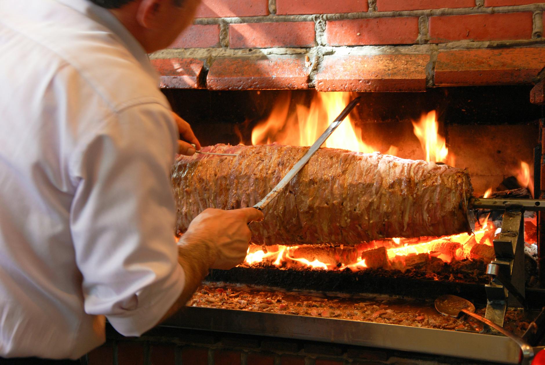 A horizontal spit of layered, wound lamb roasting beside glowing wood coals in the style of Erzurum cağ kebabı