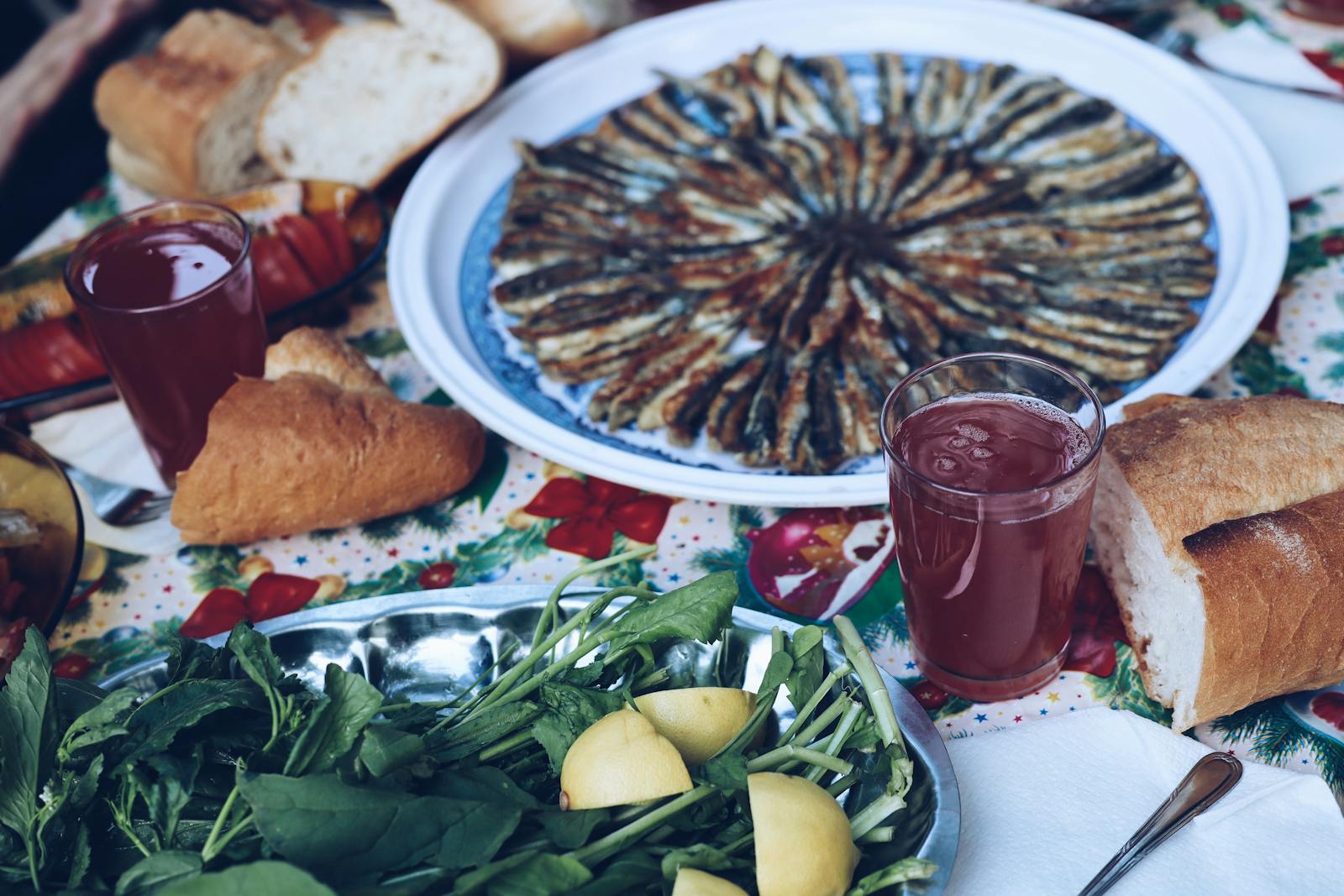 A round plate of hamsi (Black Sea anchovies) arranged in a fan, with bread, fresh greens, lemon wedges and glasses of pomegranate juice on a patterned tablecloth