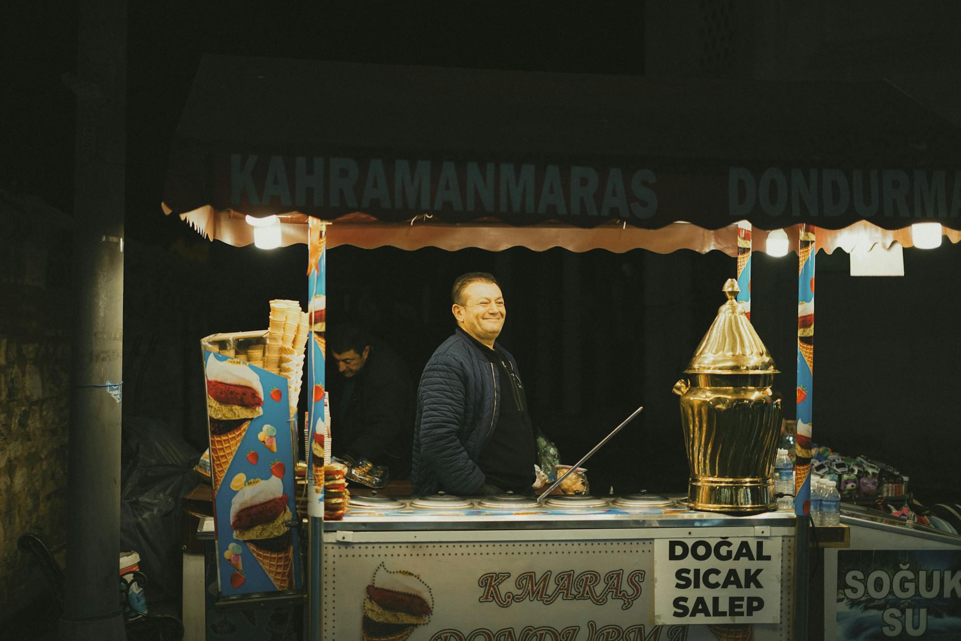 A Maraş dondurma ice cream vendor behind a lit stand at night with an ornate golden salep urn